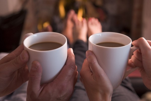 Two people holding coffee mugs in front of a fireplace. 