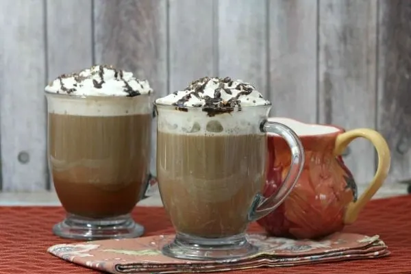 Two glasses of mocha coffee with cookie crumble topping next to a creamer mug. 