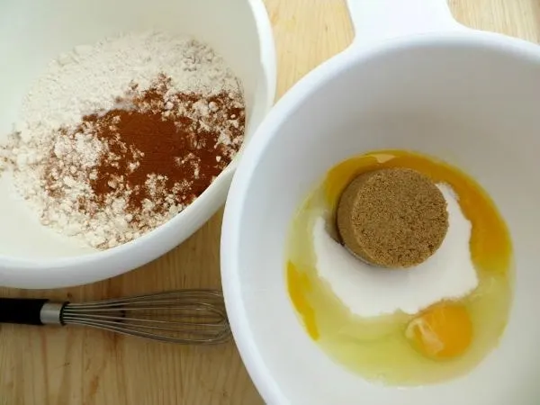 Flour and spices in a bowl next to a bowl with egg, brown sugar, and white sugar. 