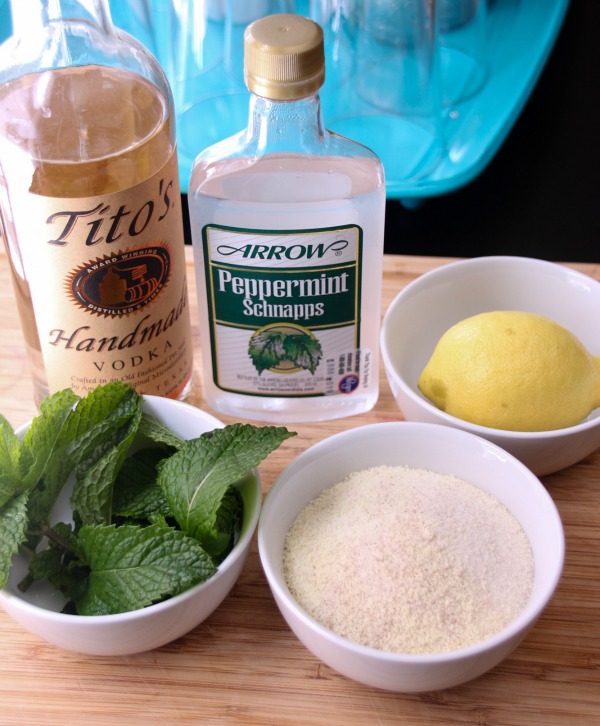 Tito's, peppermint schnapps, mint leaves, lemon, and sugar in a bowl. 