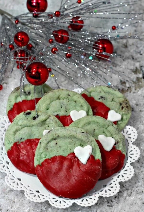 Mint chocolate chip Grinch cookies on a plate next to a silver tree with red ornaments. 