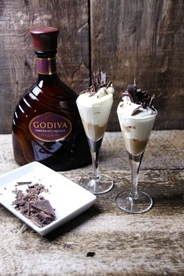 Two chocolate drinks next to a bottle of Godiva Liqueur and chocolate shavings. 