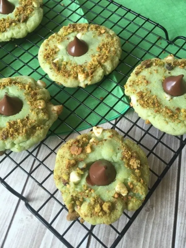 Pistachio cookies with crushed pistachios, chocolate kisses, and white chocolate chips on a cooling rack. 