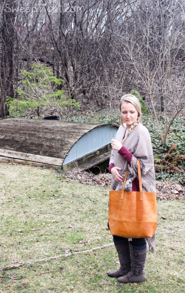 Woman standing in a yard by a bridge wearing a scarf and a tan bag. 