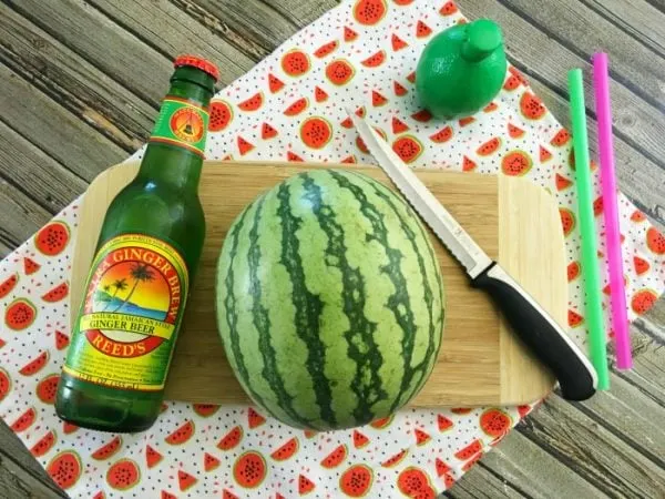 Ginger beer, watermelon, and a knife on a cutting board. 