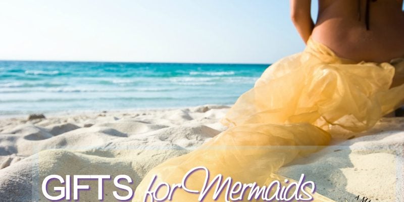 A girl that looks like a mermaid sitting in the sand by the ocean.