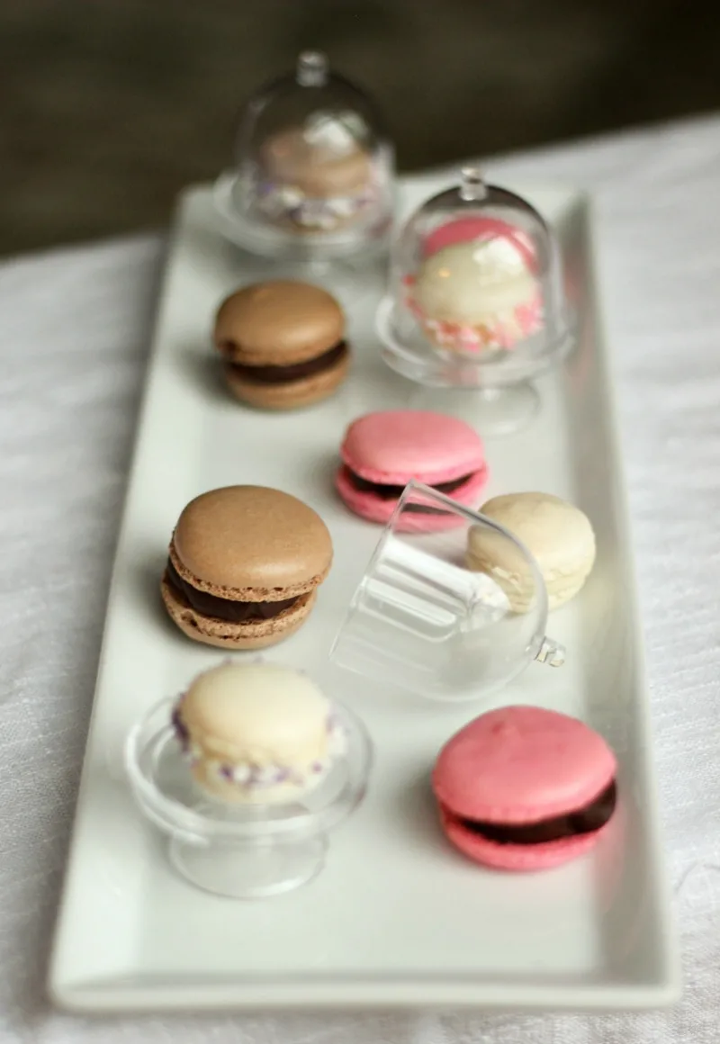 Mini macarons in brown, cream, and pink, on mini cake stands. 