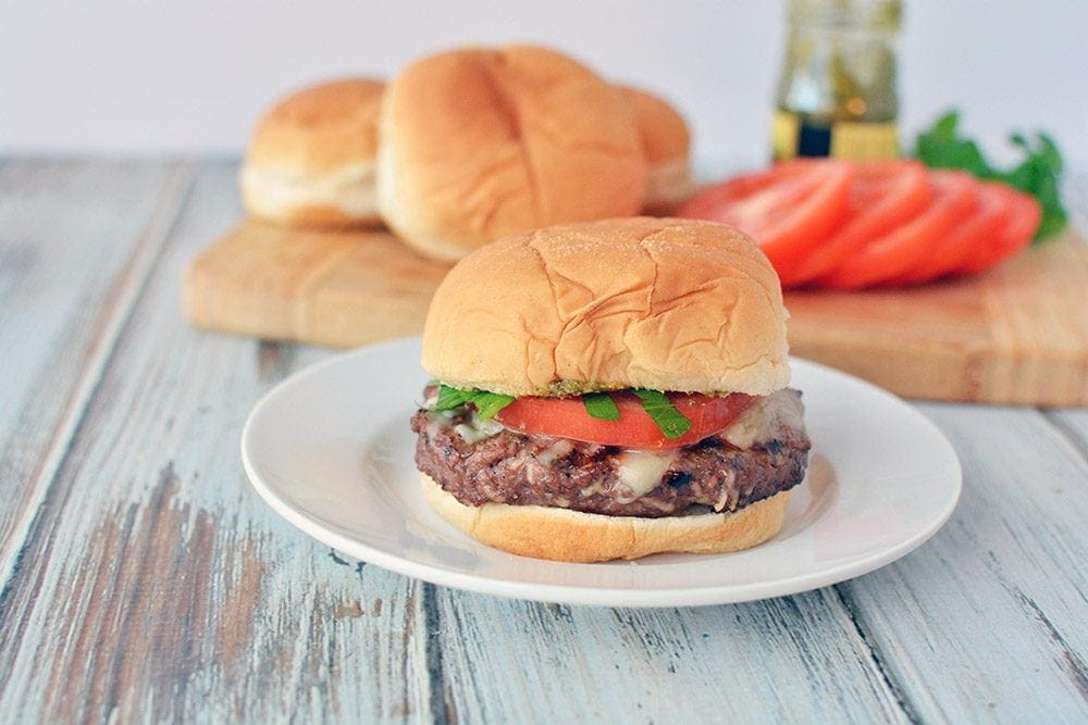 Grilled Italian burger on a plate with tomatoes and buns in the background.