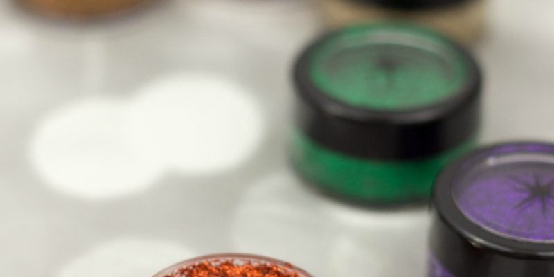 A container of orange glittery eyeshadow next to jars of green and purple.