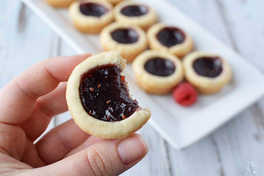 Holding a raspberry tart with a bite taken out with a tray of them in the background.