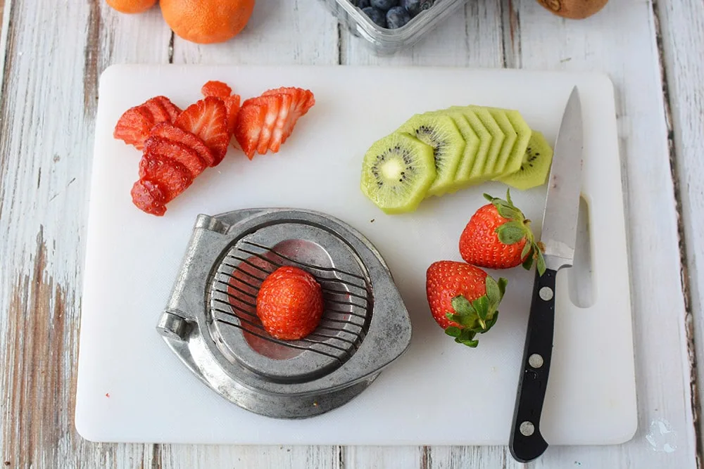 Sliced strawberries and kiwis on a cutting board with a knife and a fruit slicer. 