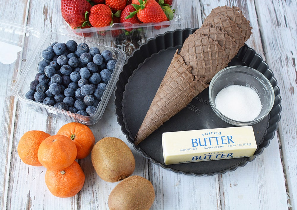 Strawberries, blueberries, kiwis, oranges, and sugar cones on a table with a tart pan and butter. 