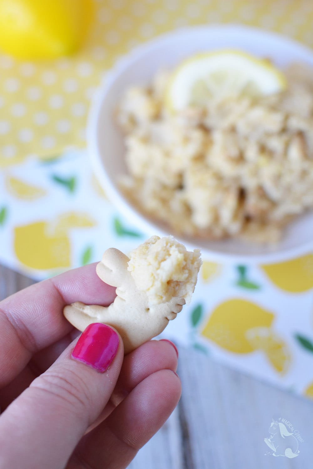 Holding an animal cracker with cookie dough on it with the bowl of cookie dough dip in the background. 