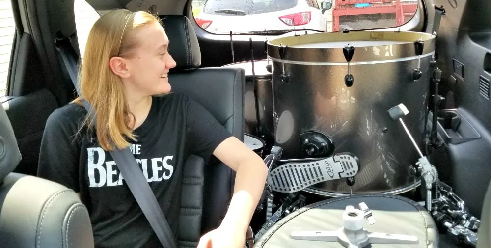 A drum set in a car with a happy teen wearing a Beatles shirt. 