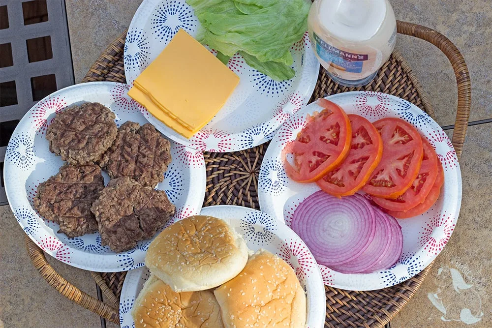 Plates of burger patties, cheese, lettuce, tomato slices, onions, and buns on a tray outside. 