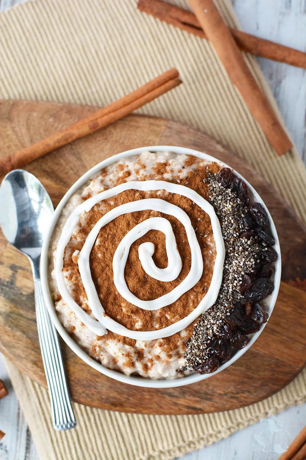 Overhead shot of smoothie bowl surrounded by cinnamon sticks. Toppings include chia seeds, raisins, and a yogurt swirl.