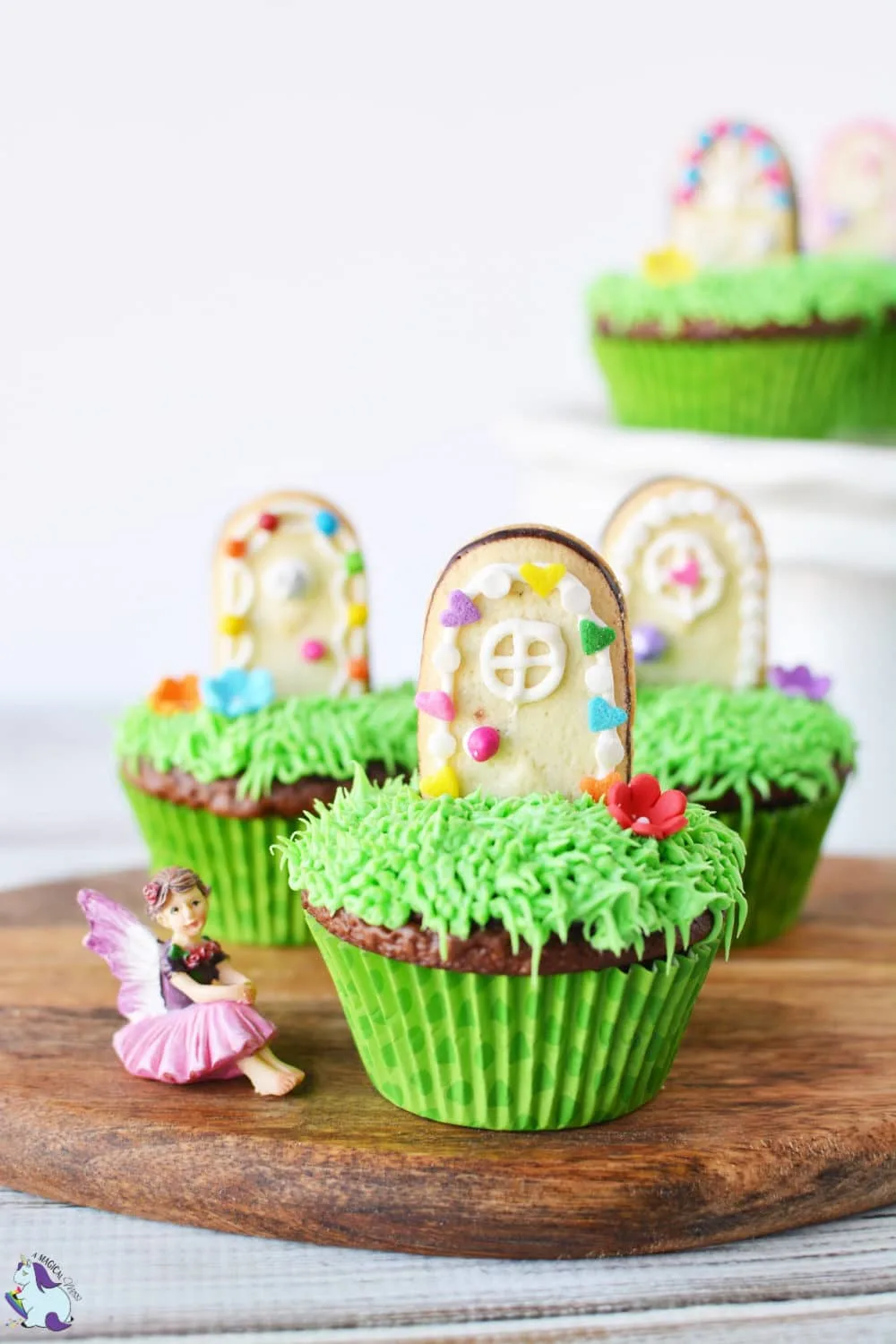 Cupcakes with green frosting and cookies decorated as doors. There is a fairy figuring sitting next to the 3 cupcakes on a cutting board. 