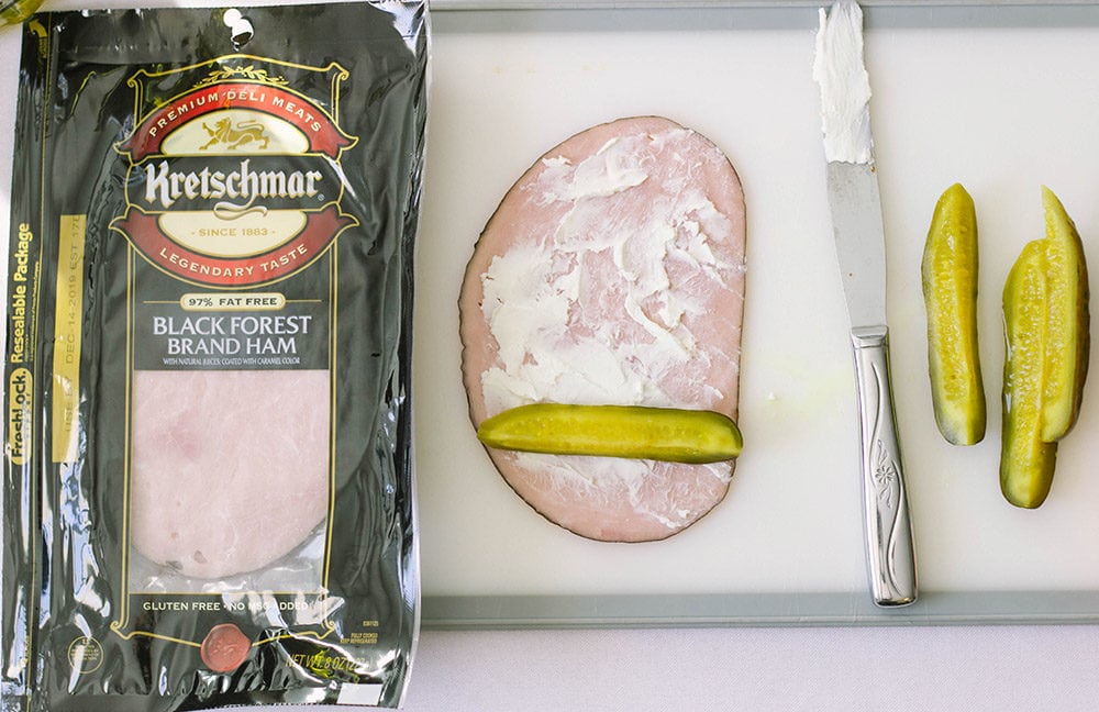 Package of ham, pickles and cream cheese on a slice of ham on a cutting board. 