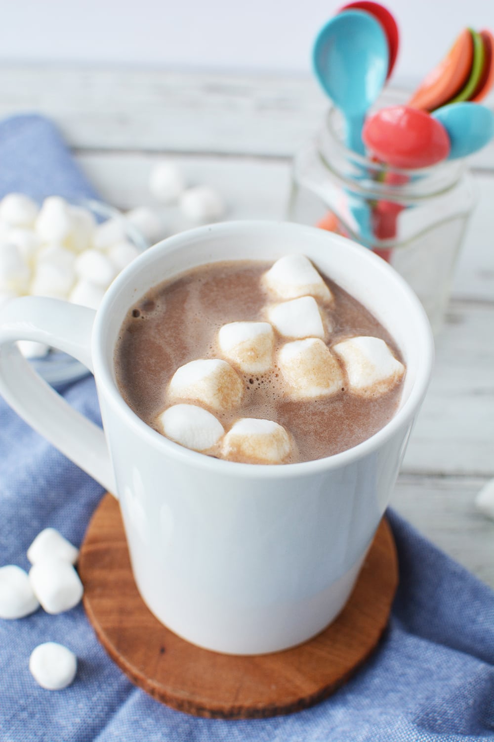 Mug of hot cocoa topped with marshmallows on a table with colored spoons in a jar. 