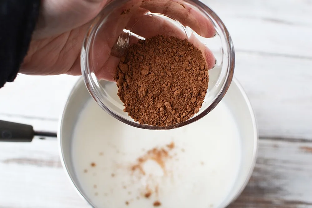 Holding a small bowl of cocoa, pouring it into a sauce pan with milk. 
