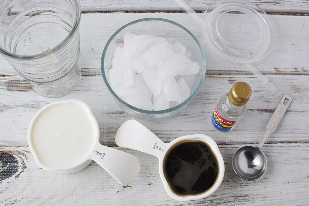 Clear tumbler, ice, milk, coffee, and flavoring on a table. 