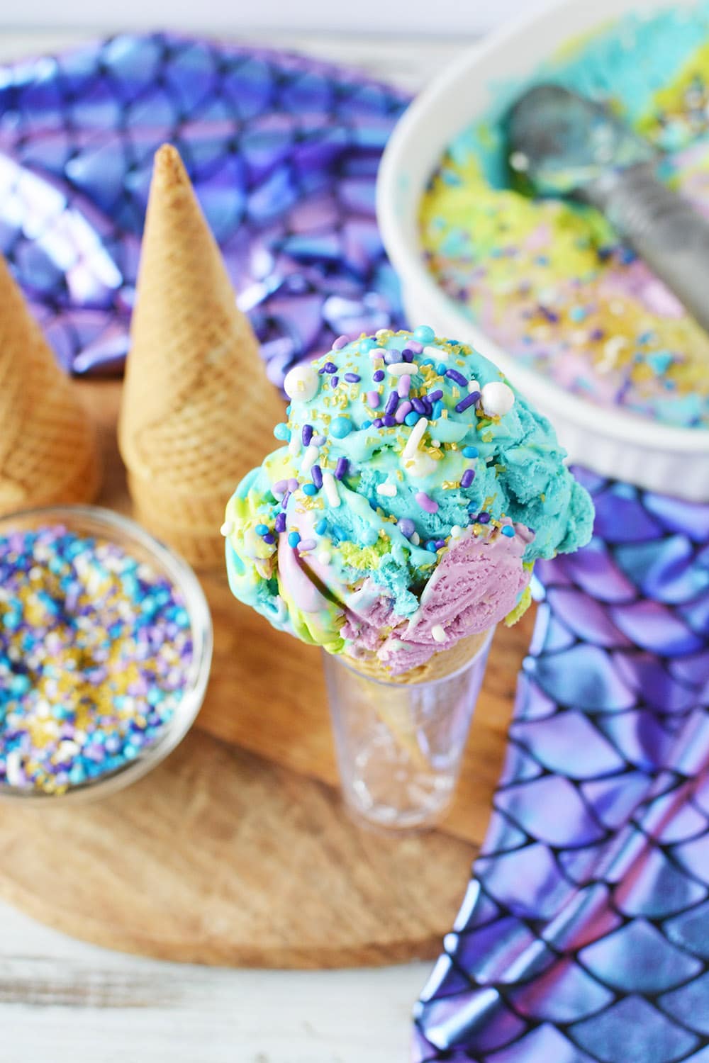 Sprinkles on blue, green, and purple ice cream with cones and dish in the background. 