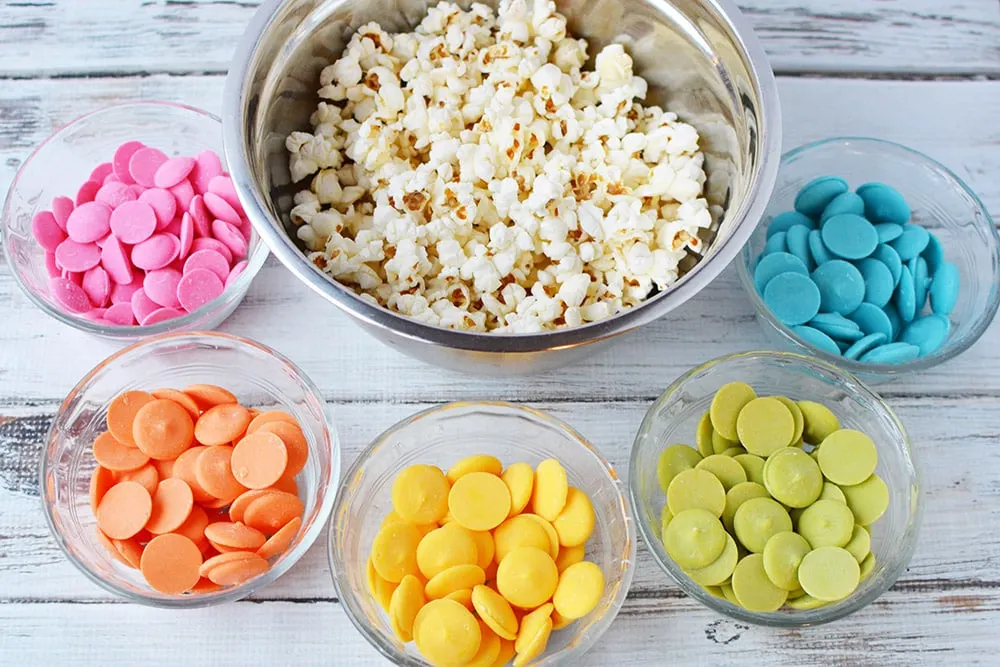 A bowl of popcorn and a rainbow of colors of candy melts in bowls. 