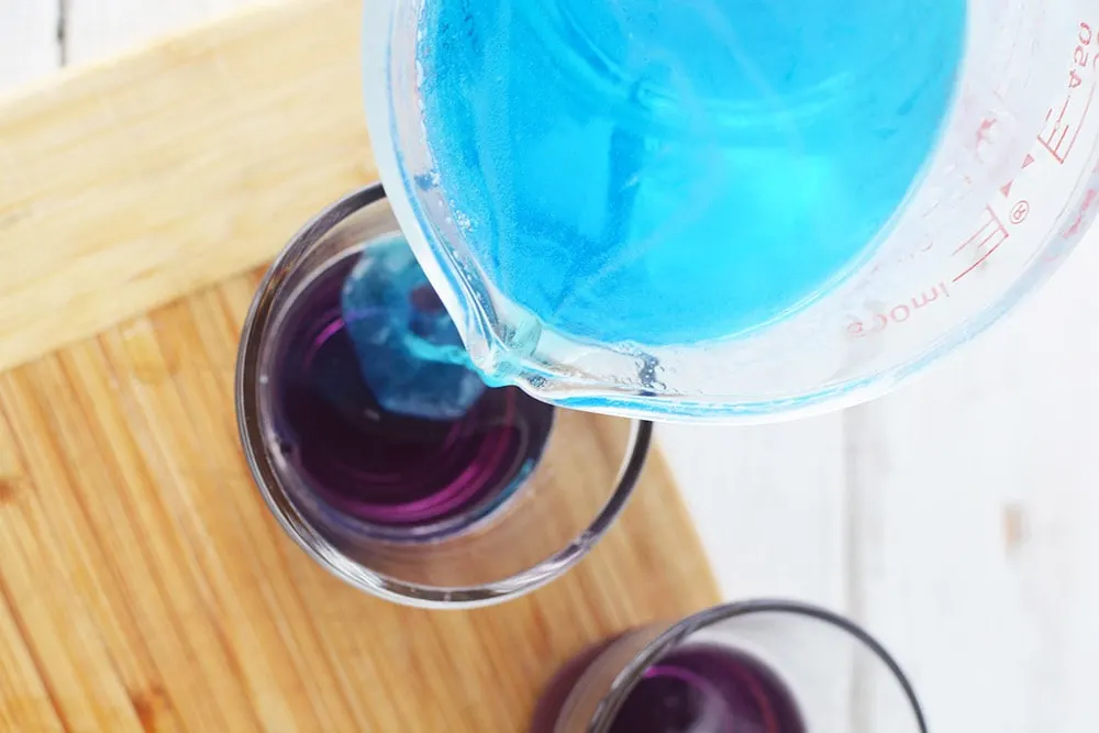 Pouring blue jello into a cup on top of purple jello. 