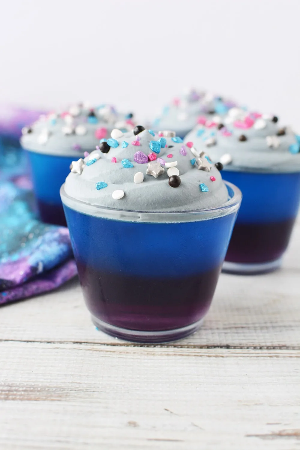Purple and blue jello cups topped with gray whipped topping and sprinkles on a table. 