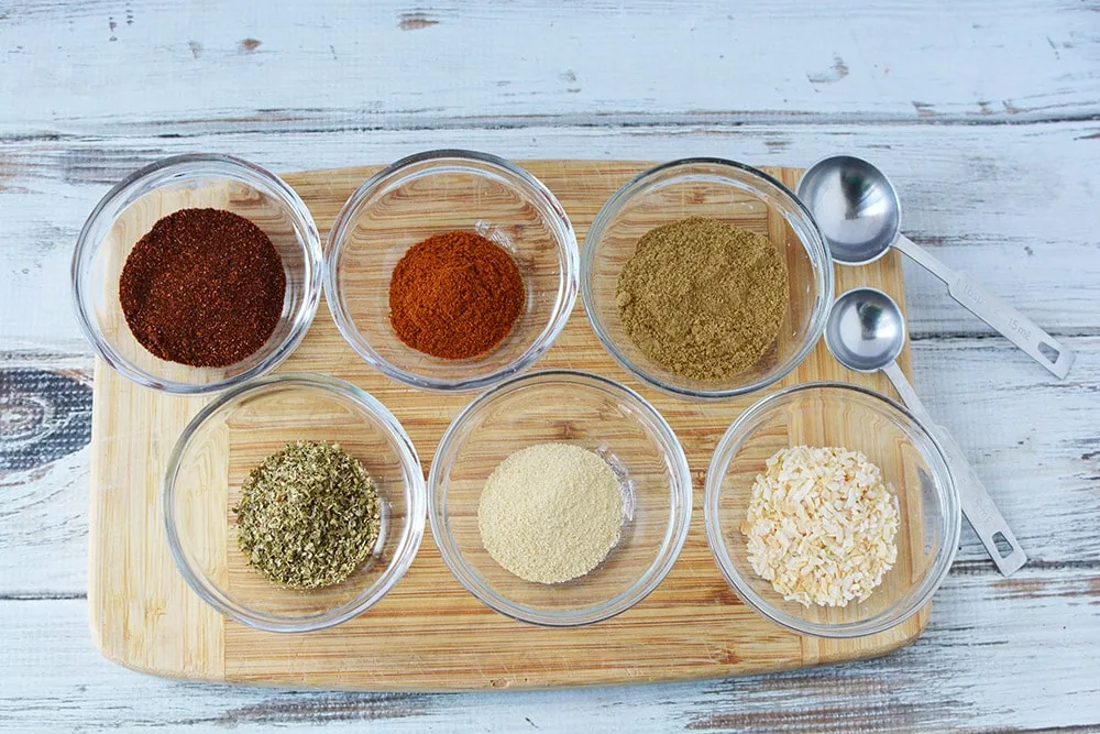 Bowls of spices for taco seasoning mix on a board next to measuring spoons. 