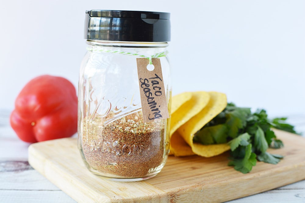 A jar of homemade taco seasoning mix on a board with taco shells and a tomato. 