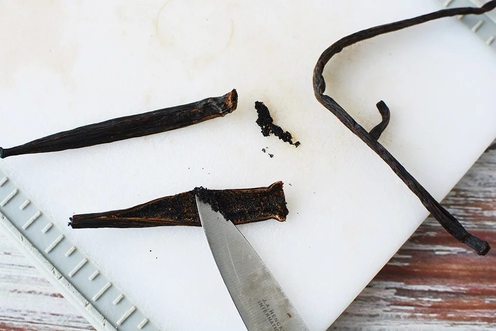 Scooping out vanilla beans with a knife on a cutting board.