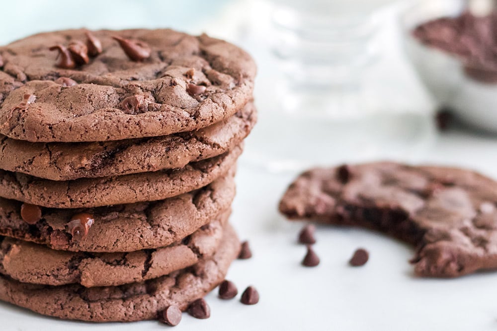 A stack of brownie cookies with a cookie missing a bite next to chocolate chips on a white table.