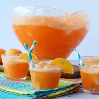 Cups of orange punch in front of large punch bowl filled with orange sherbet punch.