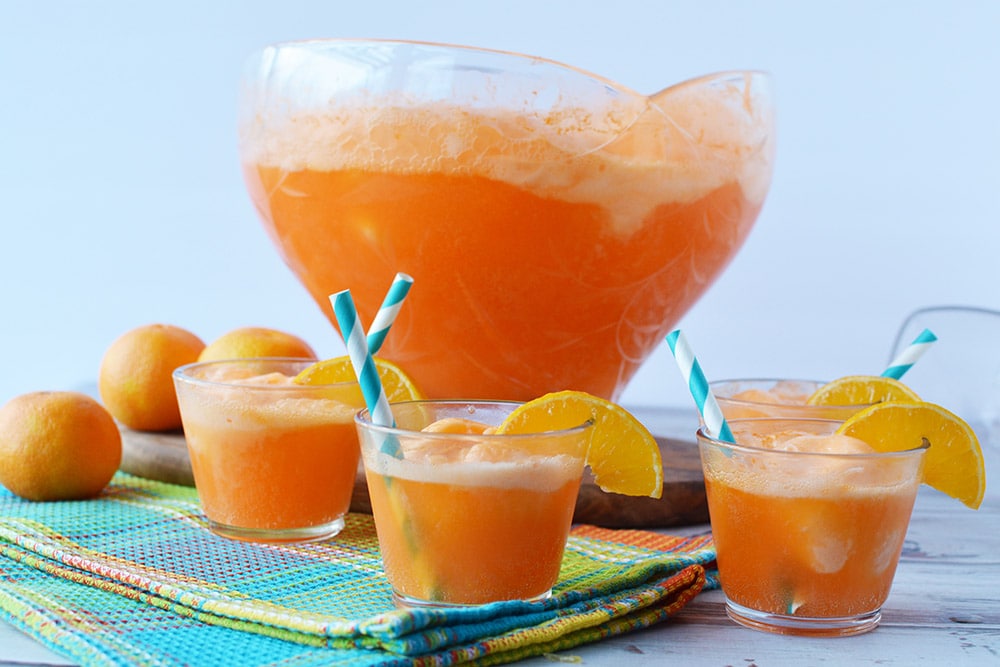 Cups of orange punch in front of large punch bowl filled with orange sherbet punch. 