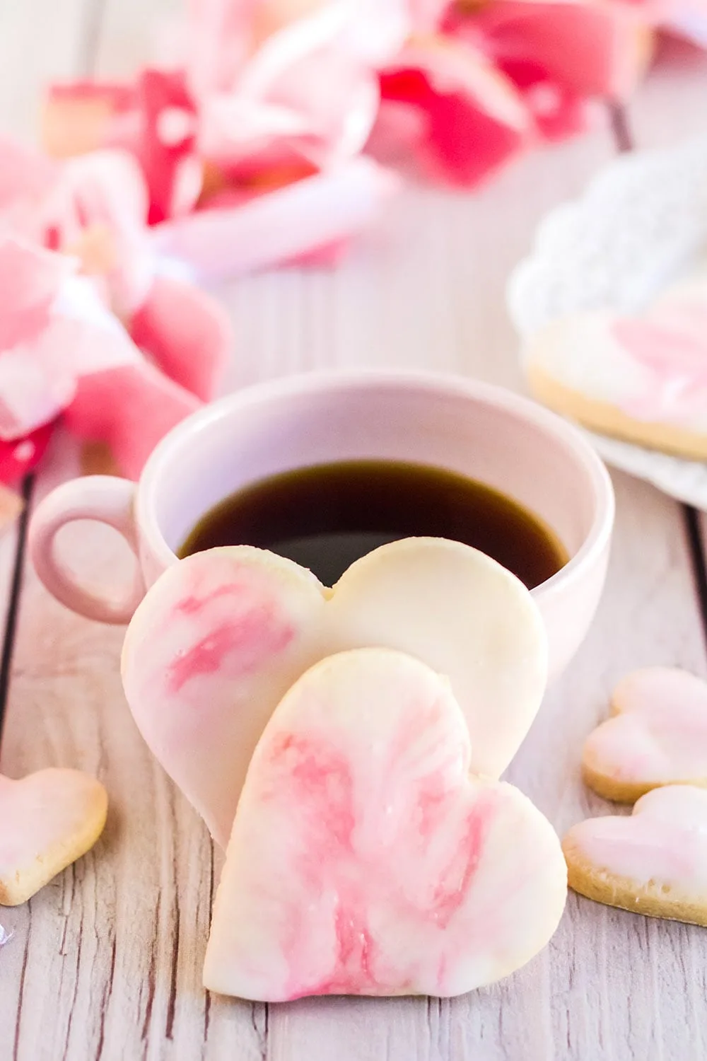 Heart cookies in front of a pink mug with coffee in it on a table with pink flowers.