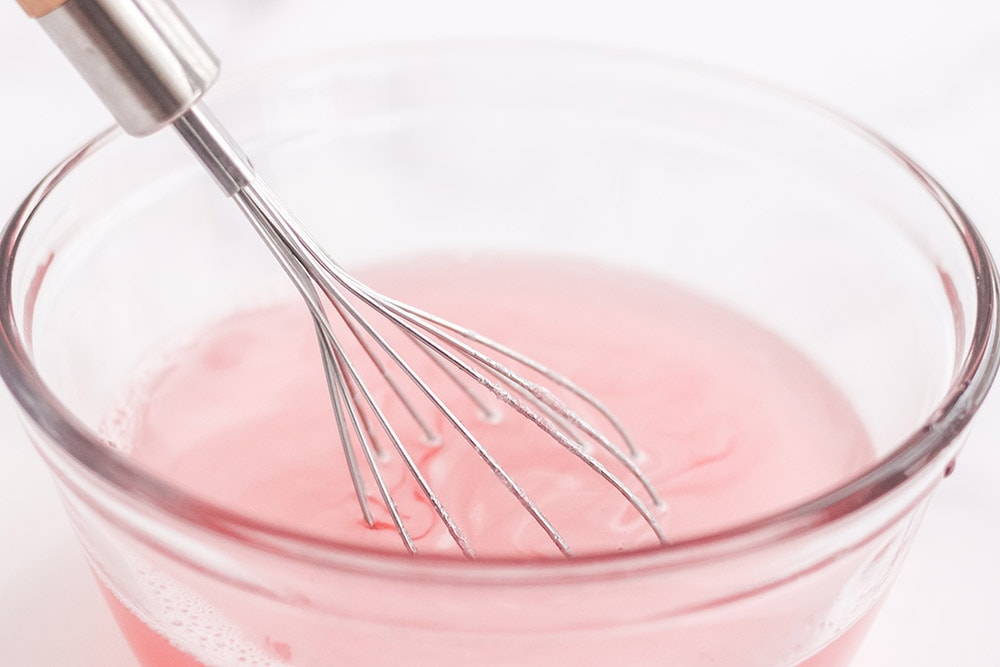 Stirring pink jello in a bowl with a whisk.
