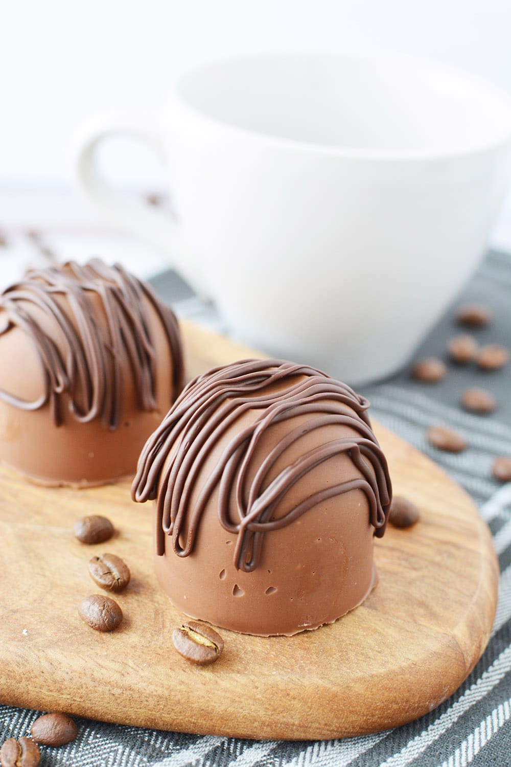 Two chocolate coffee bombs sitting on a board by a white mug.