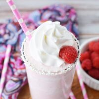 Tilted glass of raspberry frappe next to pink straws and fresh raspberries.