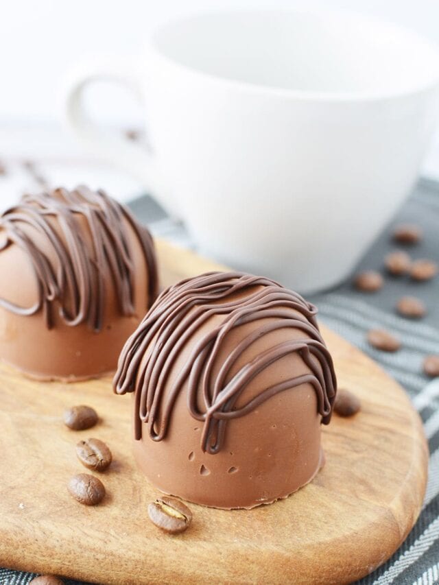 Two chocolate coffee bombs sitting on a board by a white mug.