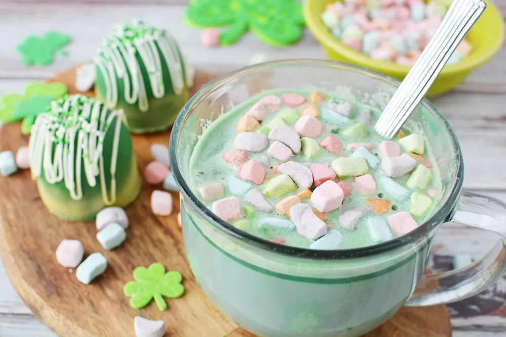 Mug of green hot chocolate with rainbow marshmallows sitting on a board next to green hot chocolate bombs and more marshmallows.