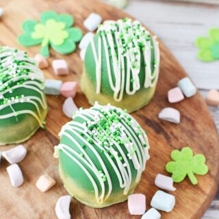 Green chocolate bombs sitting on a board with rainbow marshmallows and St. Patrick's Day decorations.