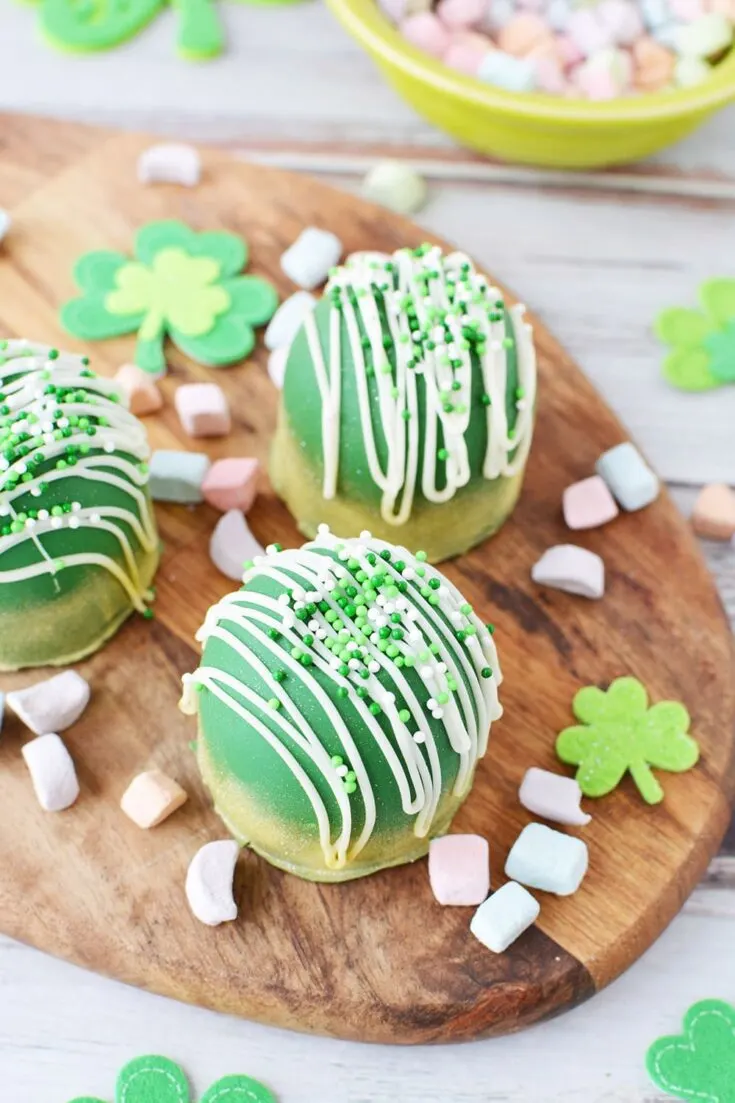 Green chocolate bombs sitting on a board with rainbow marshmallows and St. Patrick's Day decorations.