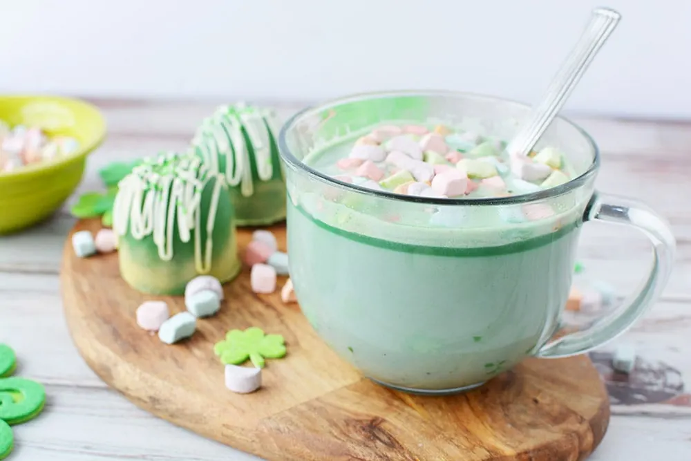 Clear mug with green hot cocoa topped with rainbow marshmallows sitting on a board next to hot cocoa bombs.