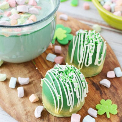 Mug filled with green hot chocolate with rainbow marshmallows next to cocoa bombs and sprinkles.