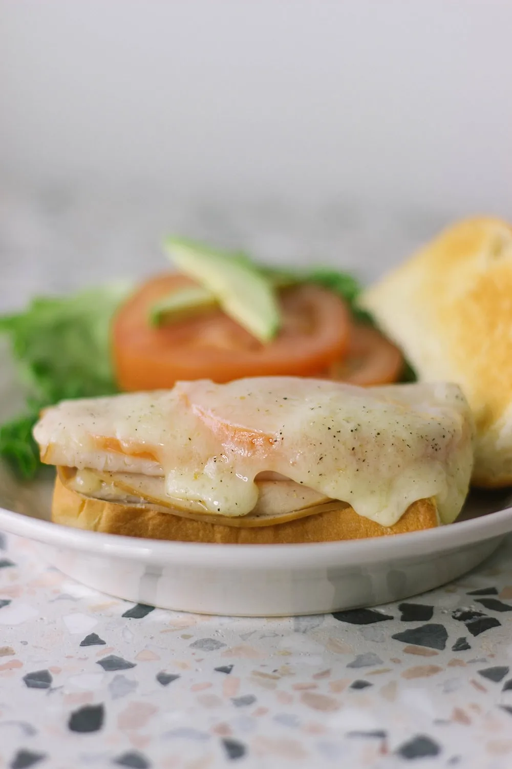 Melted cheese on a turkey melt with lettuce, tomato, and avocado in the background.