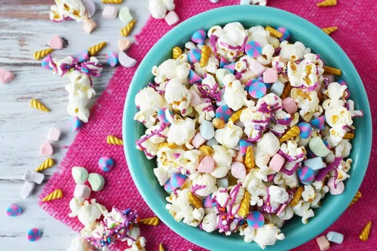 Blue bowl full of unicorn popcorn with popcorn and candies on table with a pink napkin.