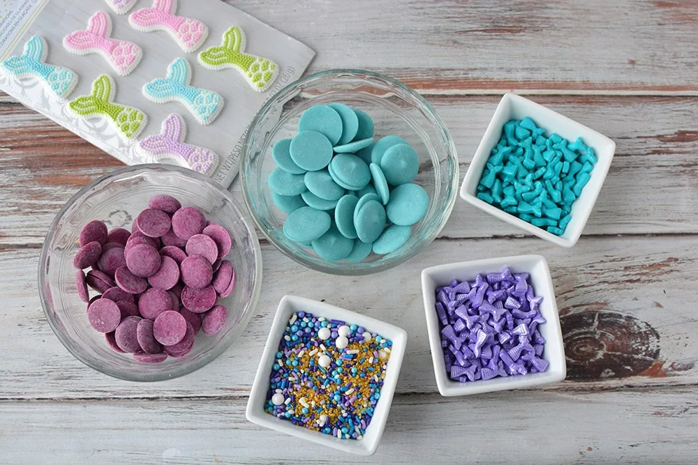 Candy fins, purple and blue melting chocolate discs, and sprinkles in bowls on a table. 