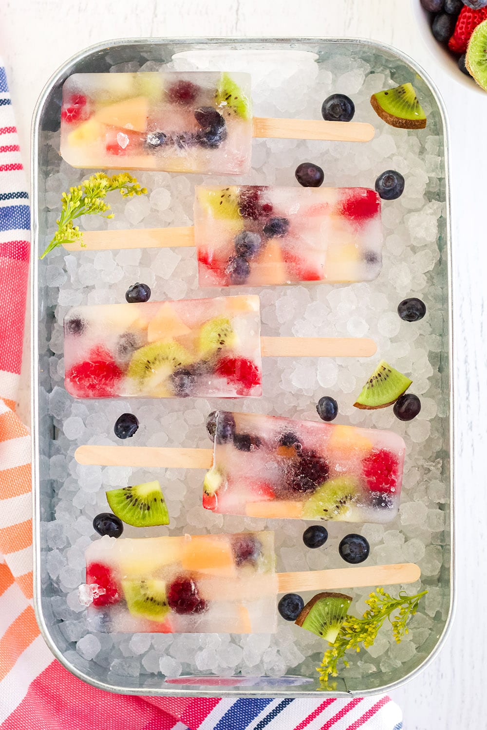 Fruit pops lined up on a bed of nugget ice with fruit in a tray.