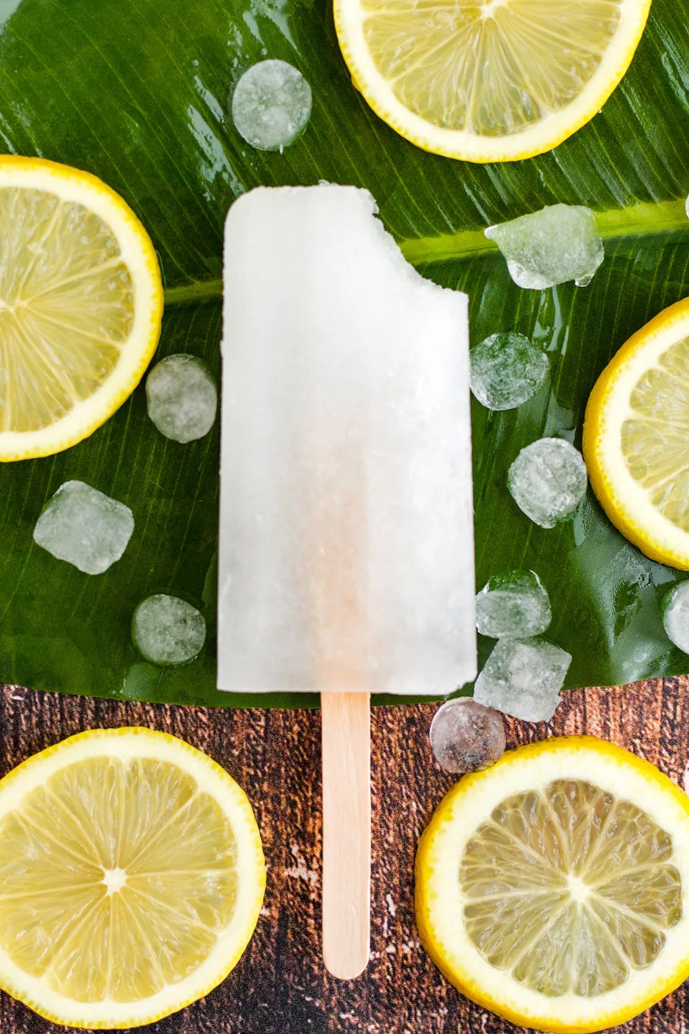 One frozen lemon pop with a bite missing with lemon slices on the table.